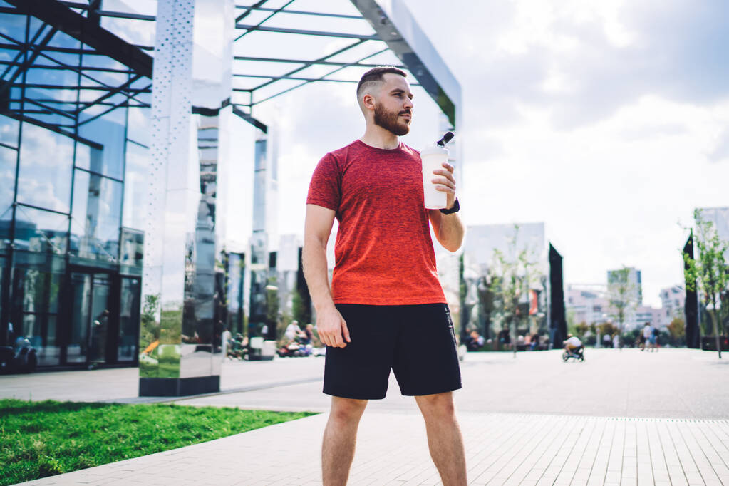 Vážný fit sportovec v aktivním oblečení stojící na chodníku v blízkosti skleněné budovy, zatímco drží .plastic láhev vody a dívá se jinam - Fotografie, Obrázek