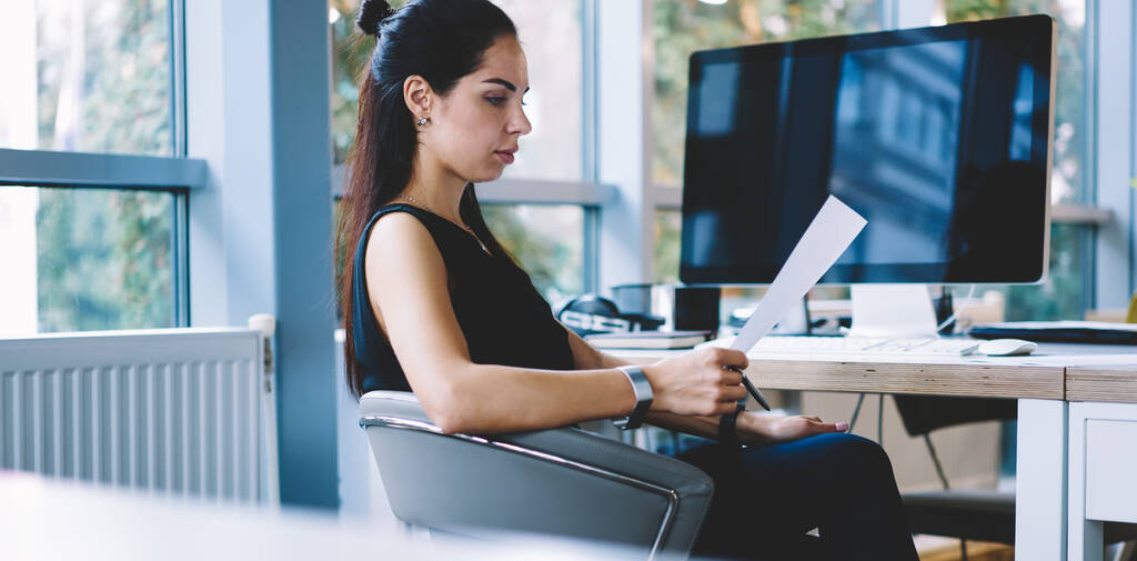 Вид збоку концентрованої бізнес-леді в стильному формальному одязі, що сидить за офісним столом з ноутбуком і читає документи вдень
 - Фото, зображення