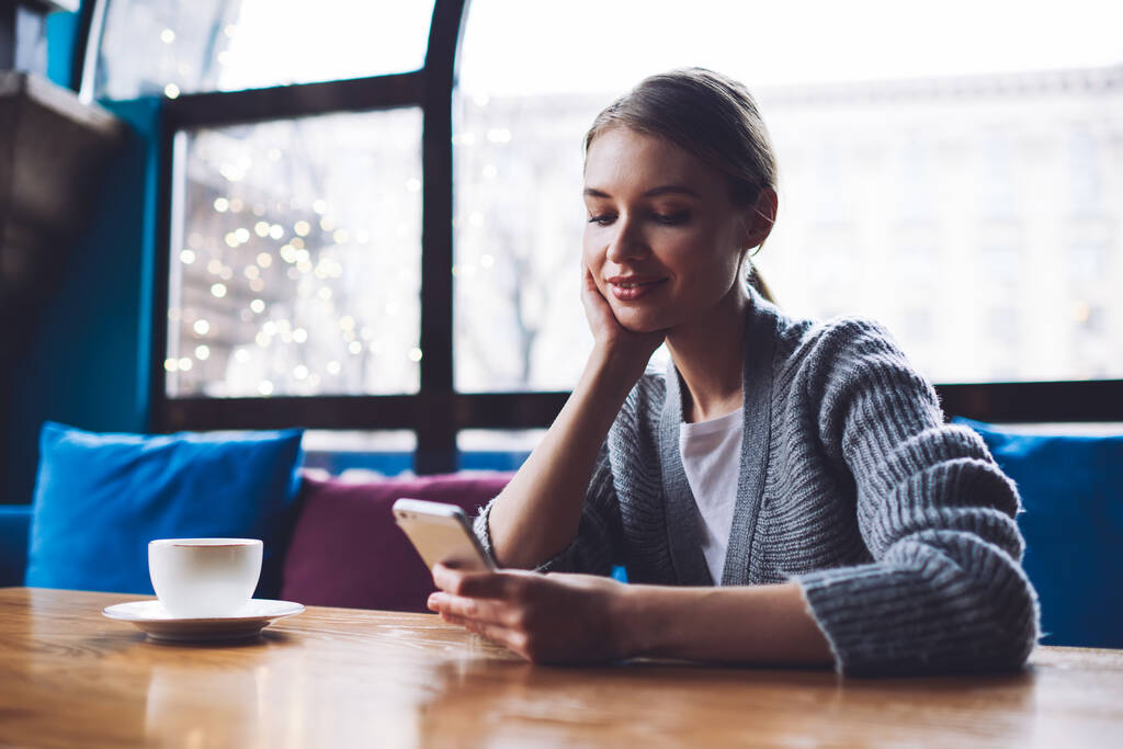 Femme souriante en vêtements décontractés assise à table avec une tasse de boisson chaude penchée sur la main et bavardant sur smartphone - Photo, image