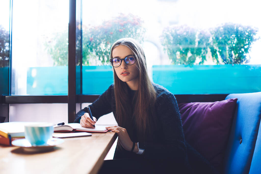 Εστιασμένη φοιτήτρια με μακριά μαλλιά και γυαλιά κοιτάζοντας την κάμερα ενώ κάθεται σε ξύλινο τραπέζι με φλιτζάνι καφέ - Φωτογραφία, εικόνα