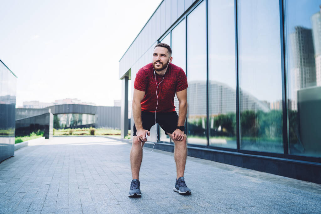 Young bearded male athlete in sportswear with earphones standing on street near building while resting after fast running and looking away - Foto, Bild