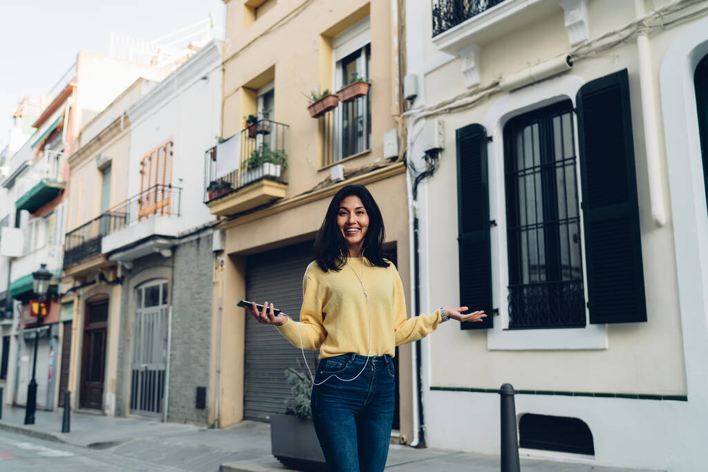 Θετική γυναίκα με casual ρούχα χαμογελώντας και περπατώντας στο στενό δρόμο ακούγοντας μουσική σε smartphone σε ακουστικά - Φωτογραφία, εικόνα