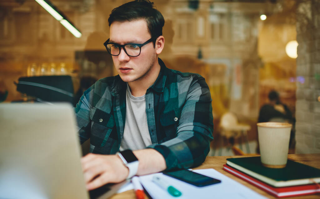 Door een glas geconcentreerde mannelijke freelancer met bril en slim horloge zittend aan tafel met een kop afhaalwarme drank browsing laptop - Foto, afbeelding