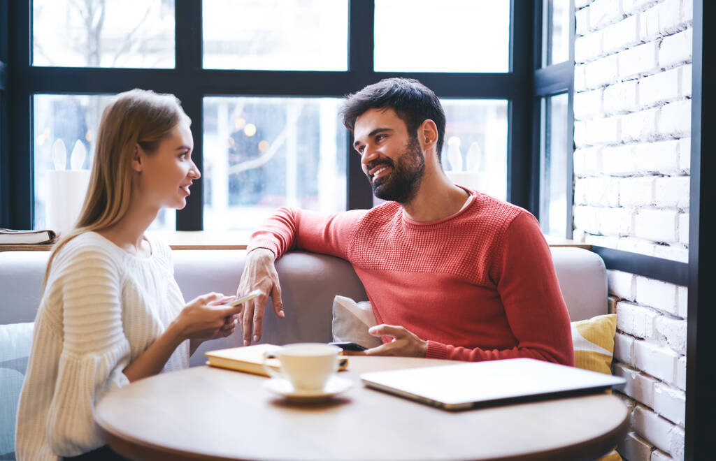 Hombre barbudo alegre en traje casual sentado con novia en suéter blanco con teléfono inteligente en cómodo sofá en la cafetería moderna - Foto, Imagen