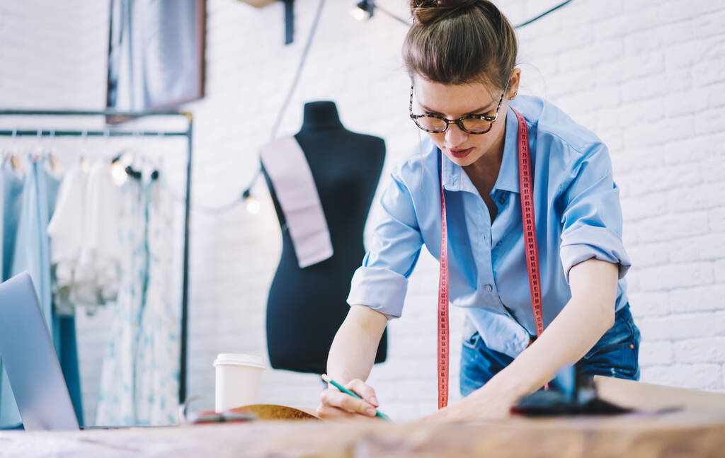 Концентрированная женщина-дизайнер в очках тонет эскиз и работает над проектом с ремесленной бумагой, стоя рядом со столом с ноутбуком в студии - Фото, изображение