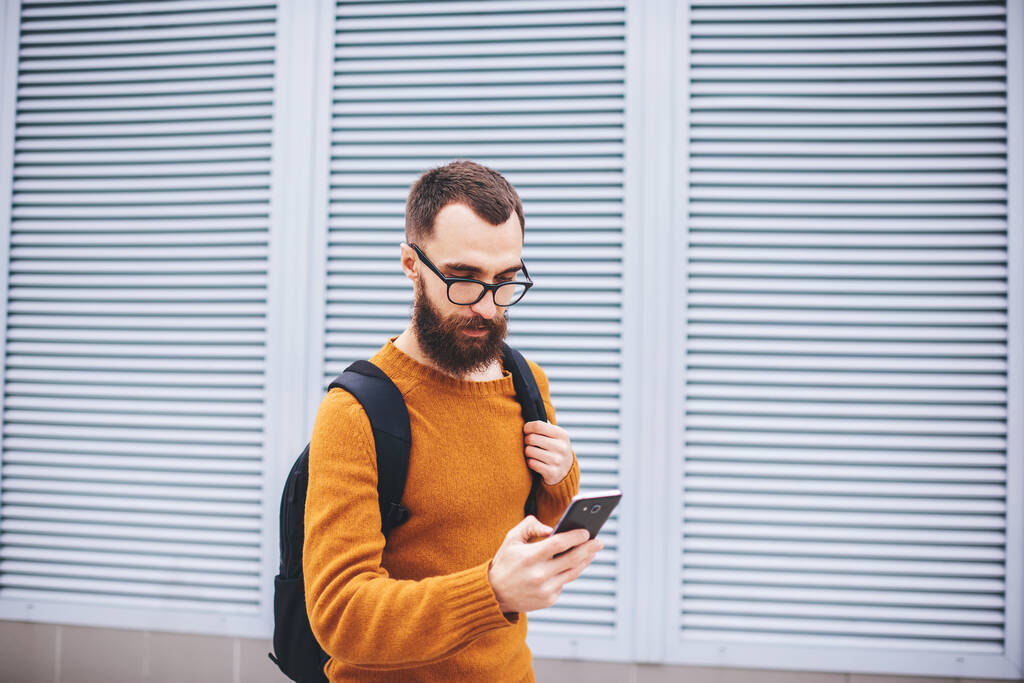 Молодий стрункий бородатий хіпстер в жовтих окулярах з чорним рюкзаком читає текстове повідомлення на мобільному телефоні біля сучасної будівлі
 - Фото, зображення