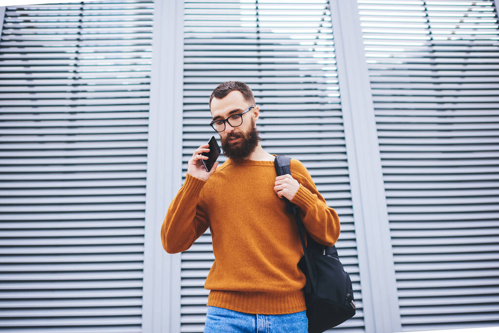 歩きながら携帯電話で会話をしながら通りに立っているバックパック付きのカジュアルな服の思慮深い男性の低角度 - 写真・画像