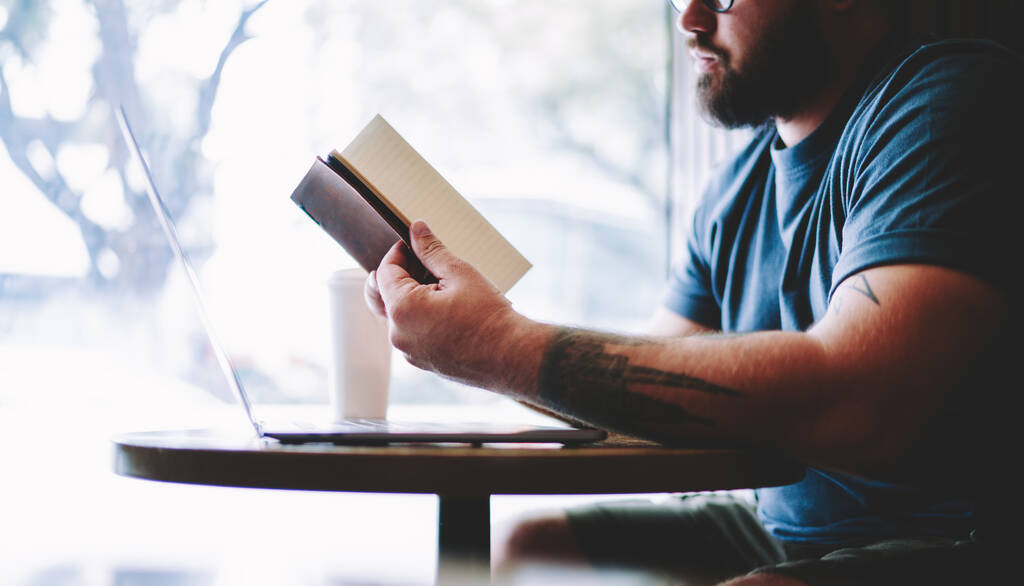 Вид збоку бородатого чоловіка, що сидить за столом з відкритими ноутбуками для читання ноутбуків у блокноті, проводячи час у кафе
 - Фото, зображення
