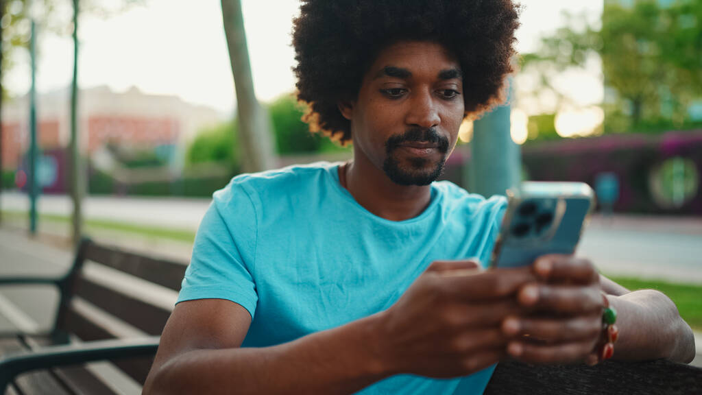 Close-up of young African American man in blue t-shirt sitting on park bench and talking on speakerphone. Smiling man sends voice message on mobile phone. lifestyle concept.  - Photo, Image