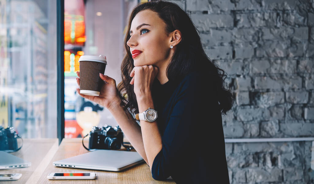 Side view of thoughtful female photographer with wristwatch standing with takeaway coffee at wooden desk with laptop and smartphone while looking away - Photo, Image