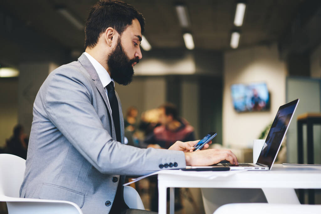Zijaanzicht van geconcentreerde ondernemer met baard in formele kleding zittend aan tafel en bladerend op laptop en smartphone tijdens het werk in kantoor - Foto, afbeelding