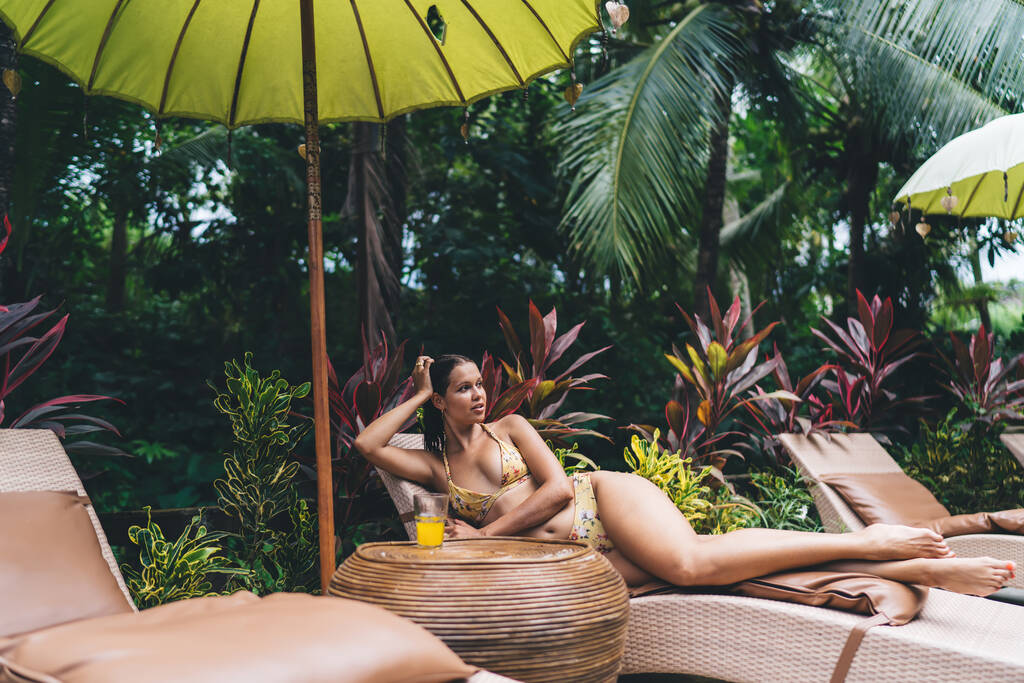 Corps complet de femme ethnique sensuelle en maillot de bain relaxant sur une chaise longue près de la piscine et bronzer pendant les vacances d'été - Photo, image