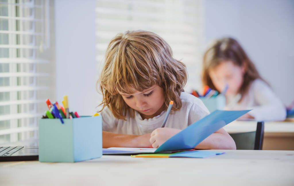 Niño de la escuela escribiendo algo en copybook y sentado en la mesa en el aula. Retrato de niño dibujando en copybook - Foto, imagen