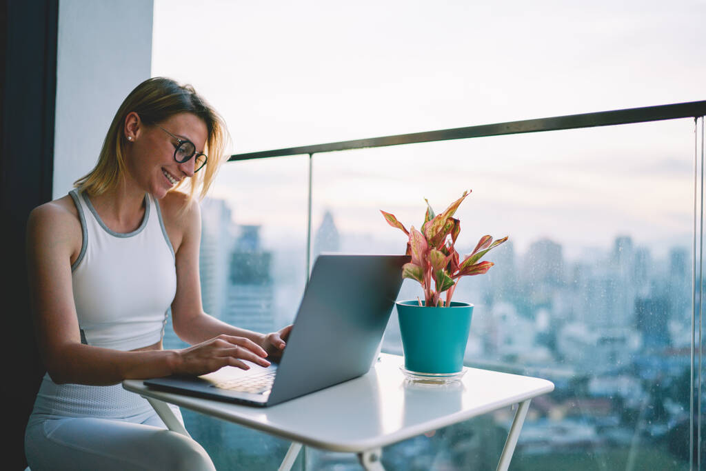 Young smiling blond female in white top and trousers typing letter on laptop sitting at small table on apartment balcony - Foto, Bild