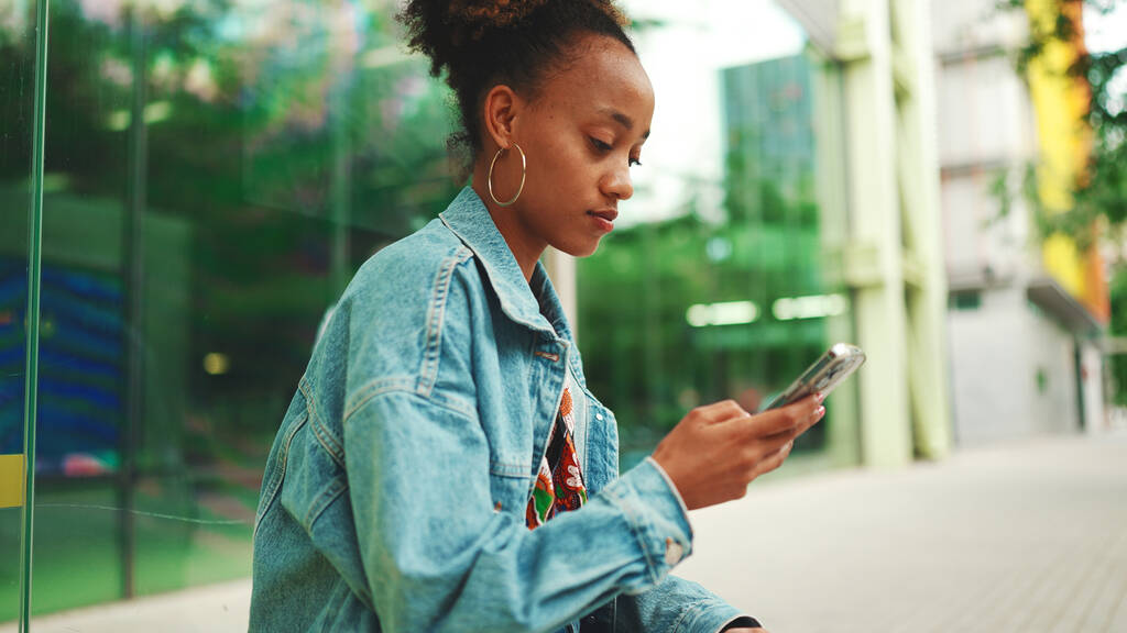 Симпатичная африканская девушка с хвостиком, в джинсовой куртке, в кукурузном топе с национальным рисунком, сидит на автобусной остановке и использует свой мобильный телефон.  - Фото, изображение