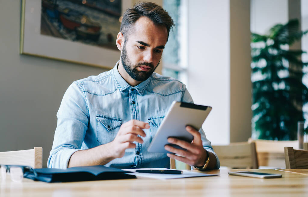 Freelancer focado sentado à mesa com notebook e documentos e tablet de navegação enquanto trabalhava no projeto no espaço de trabalho criativo durante o dia - Foto, Imagem
