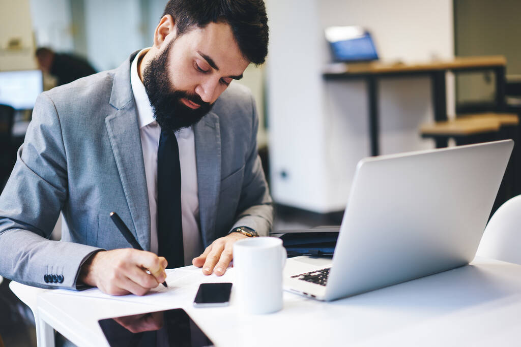 Empresário pensativo com barba em terno formal sentado à mesa com laptop e xícara de bebida e escrevendo nota na folha de papel - Foto, Imagem