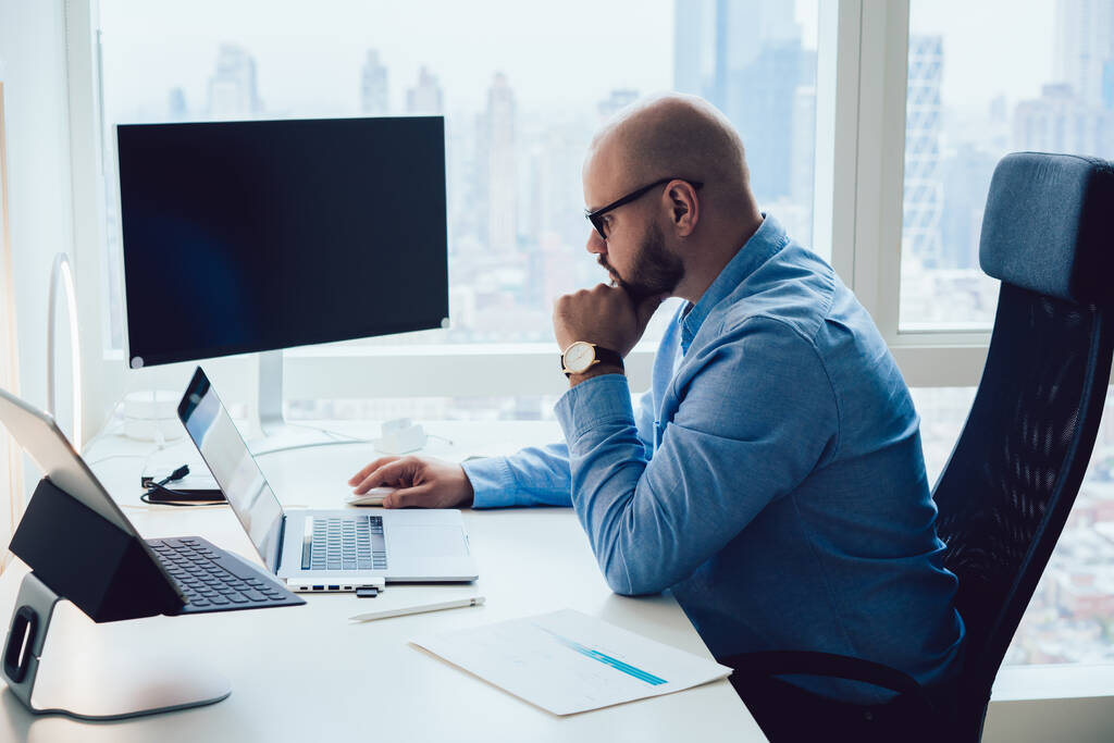Вид збоку концентрованого молодого безволосистого бородатого чоловіка-менеджера в стильному вбранні та окулярах, що працюють на ноутбуці, сидячи за столом з рукою підборідді
 - Фото, зображення