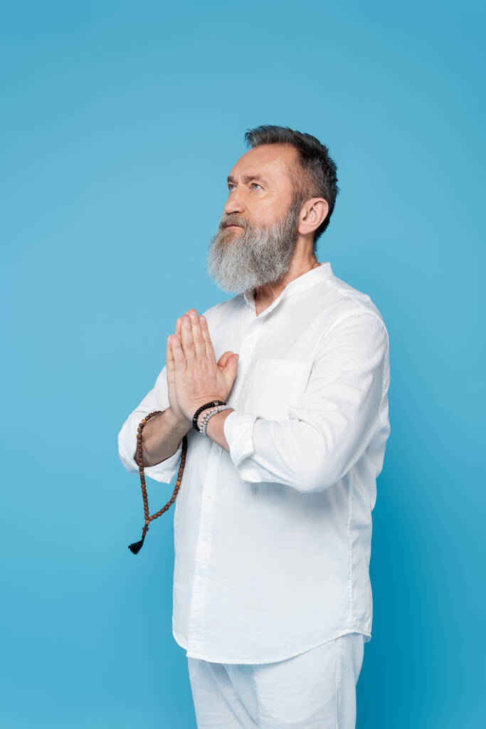 entrenador espiritual barbudo con cuentas de mala meditación con las manos orantes aisladas en azul - Foto, Imagen