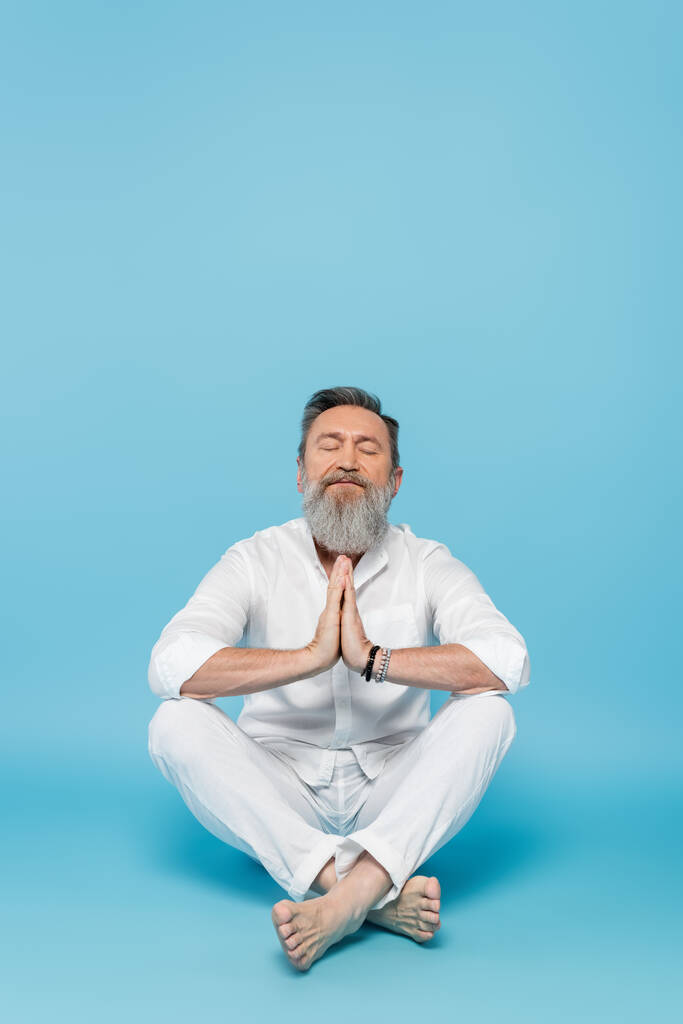 bearded man with closed eyes and praying hands meditating in easy pose on blue  - Photo, Image