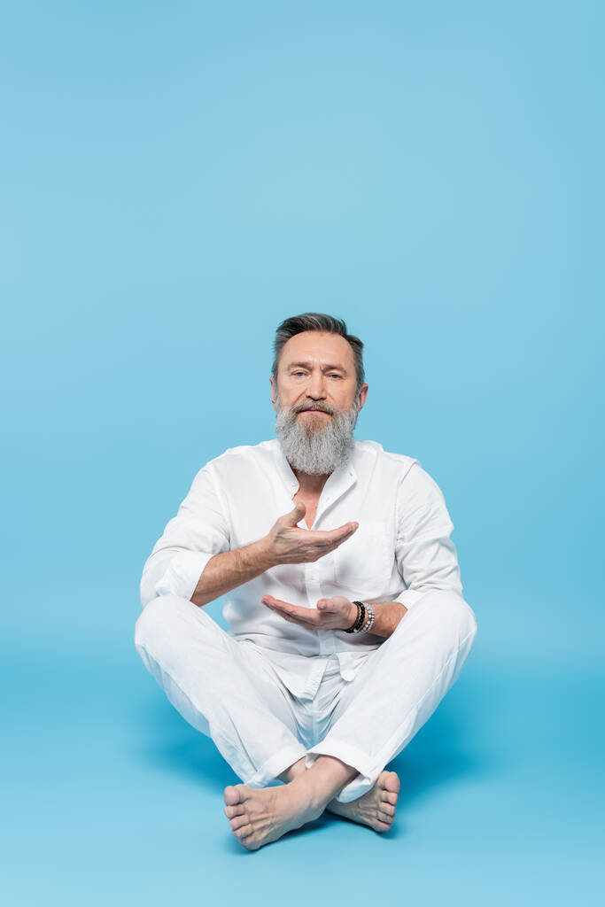 bearded man in white clothes meditating in easy pose on blue background - Photo, Image