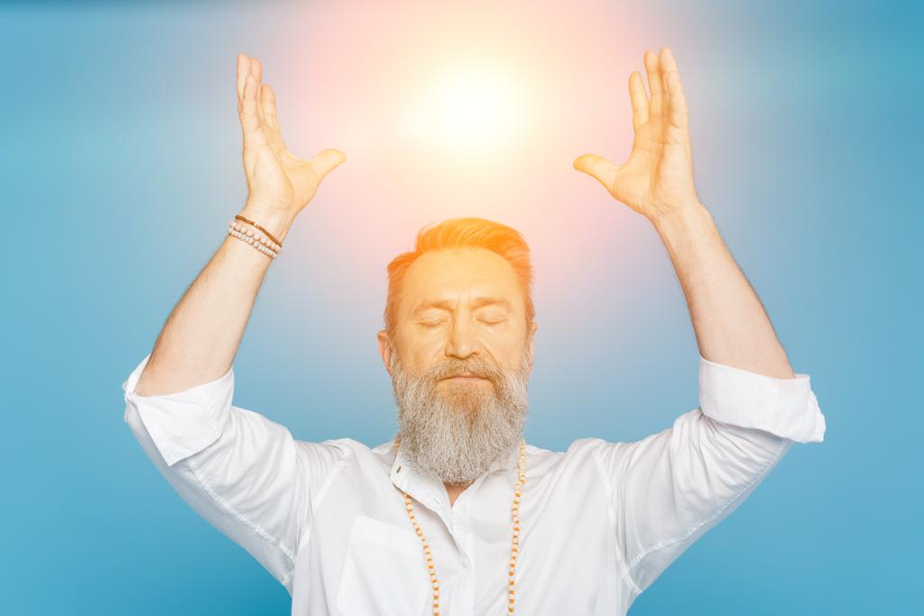 Meister-Guru meditiert mit geschlossenen Augen und erhobenen Händen in der Nähe einer leuchtenden Aura, isoliert auf blauem Grund - Foto, Bild
