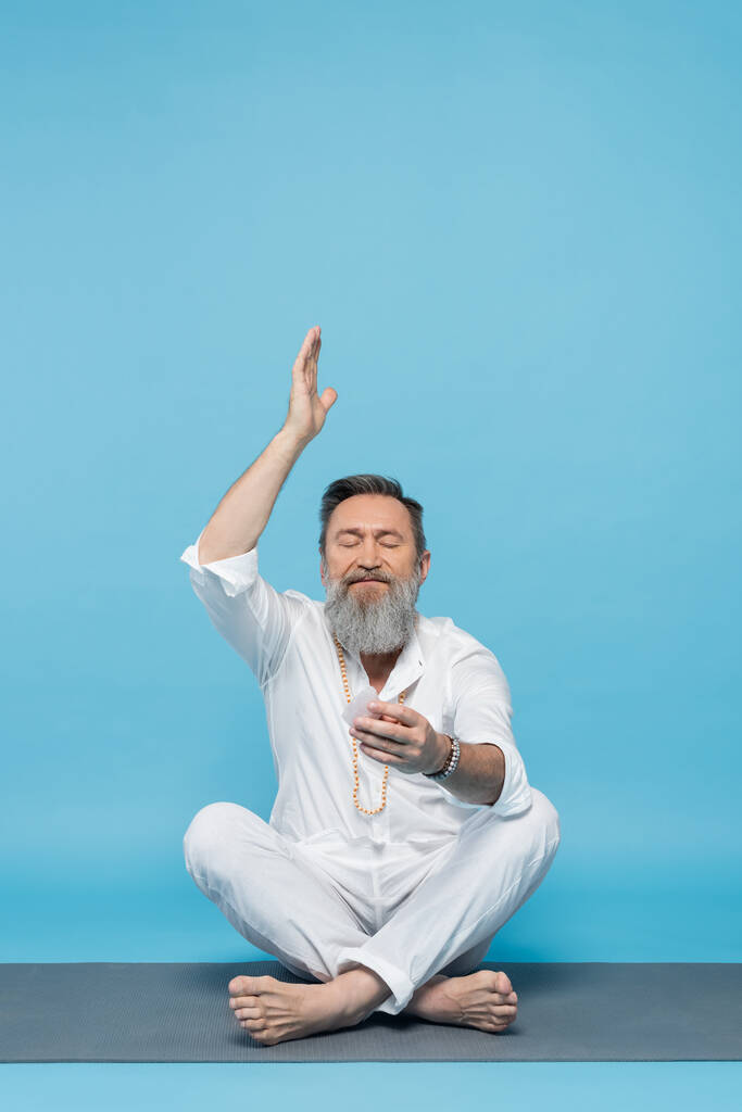 ανώτερος άνθρωπος σε λευκά ρούχα διαλογίζεται με κρύσταλλο σεληνίτη σε εύκολη στάση με έθεσε το χέρι στο μπλε - Φωτογραφία, εικόνα