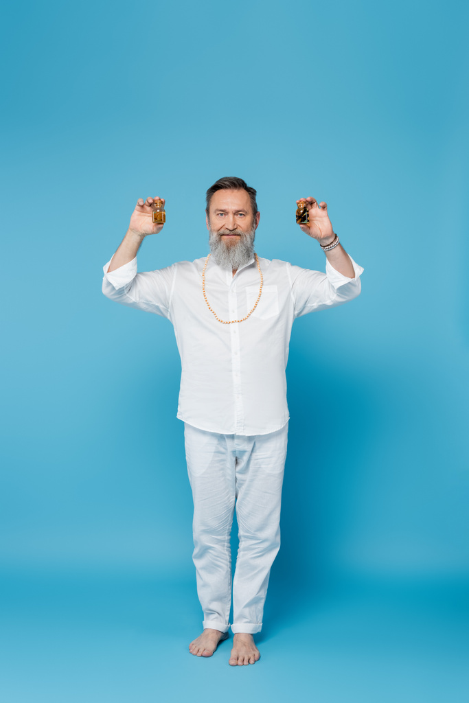 hombre barbudo descalzo en ropa blanca sosteniendo botellas con aceites esenciales en azul  - Foto, Imagen