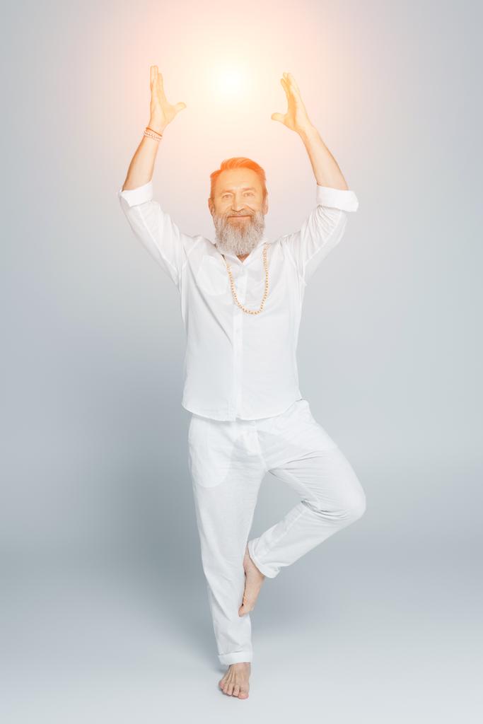 Yoga-Guru meditiert in voller Länge in Baumpose neben leuchtendem Chakra auf grau - Foto, Bild