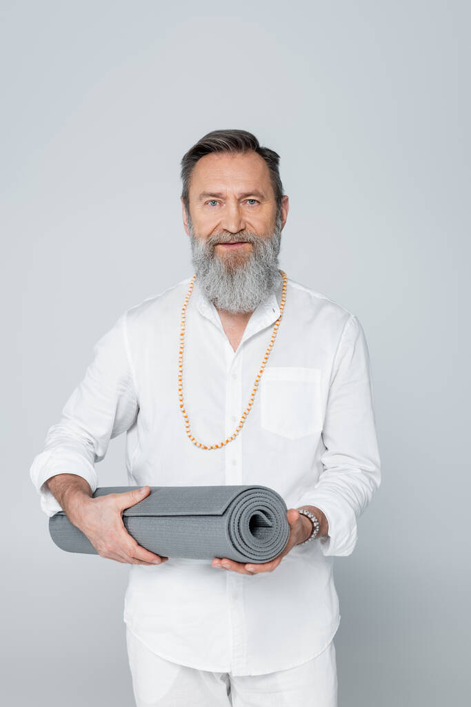 bearded master guru in white shirt and beads holding yoga mat isolated on grey - Photo, Image