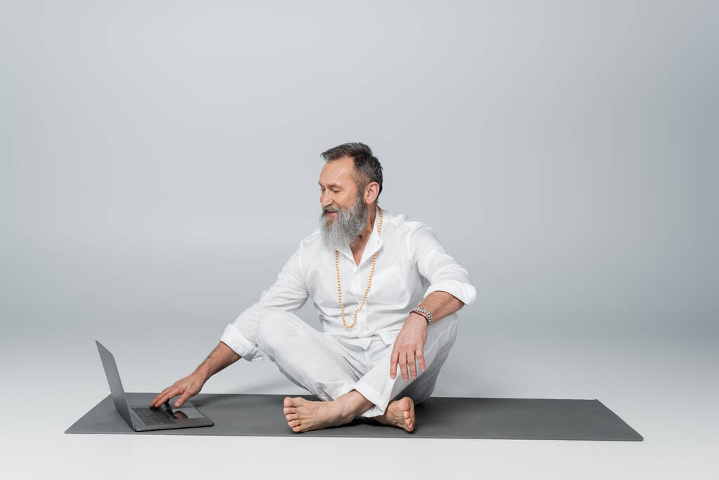 mentor spirituel souriant assis sur un tapis de yoga en pose facile près d'un ordinateur portable sur du gris - Photo, image