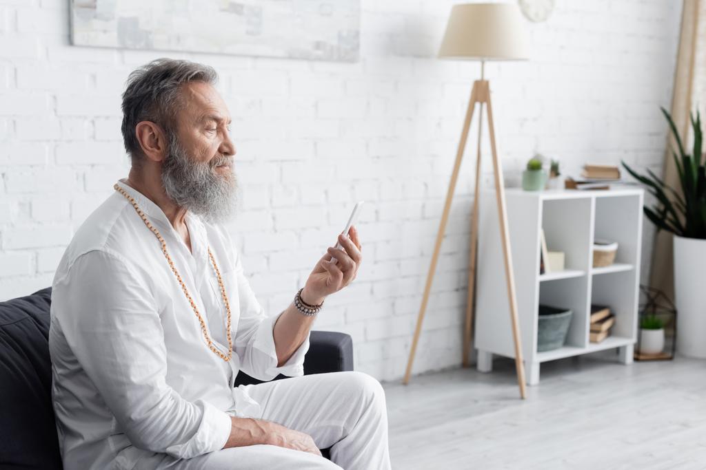 parrakas guru mies valkoisissa vaatteissa istuu sohvalla ja katselee matkapuhelinta - Valokuva, kuva