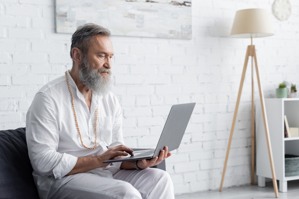 szakállas spirituális edző fehér ruhában ül a kanapén és laptopot használ - Fotó, kép