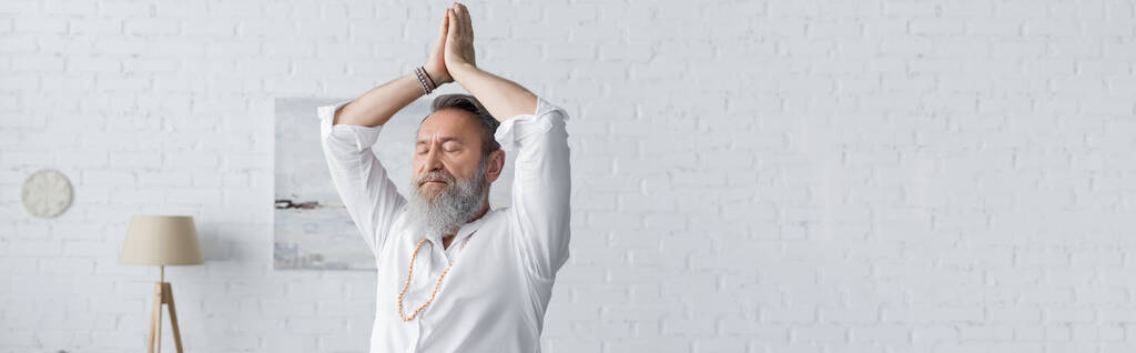 starszy brodaty mężczyzna w białej koszuli medytujący z rękami w górze i modlącymi się rękami, sztandar - Zdjęcie, obraz