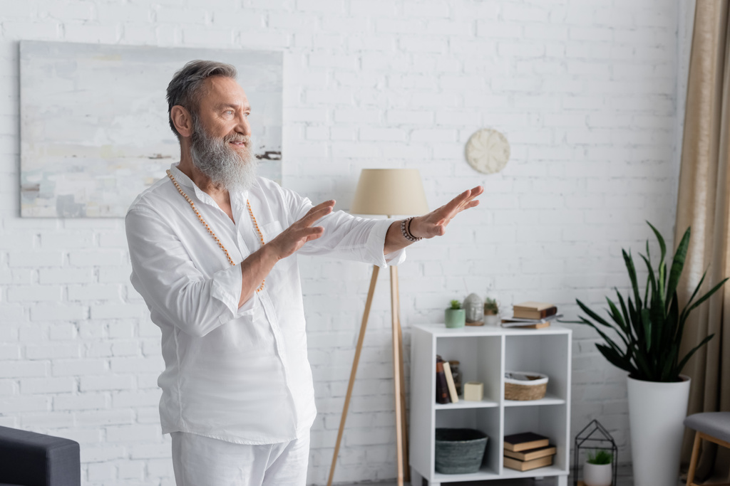 bärtiger Guru im weißen Hemd meditiert zu Hause mit ausgestreckten Händen - Foto, Bild