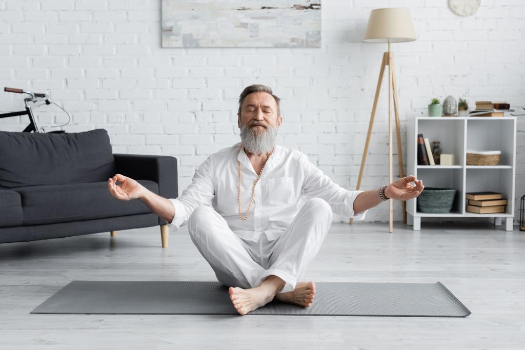Бородатый тренер-гуру медитирует в легкой позе с закрытыми глазами в гостиной - Фото, изображение