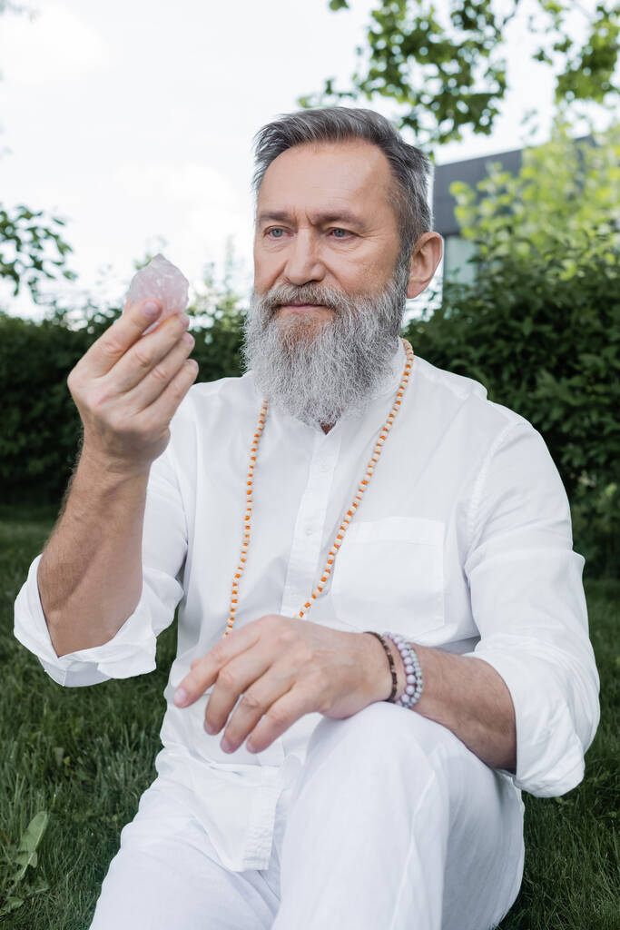 maître gourou senior en chemise blanche et perles regardant le cristal de sélénite à l'extérieur - Photo, image