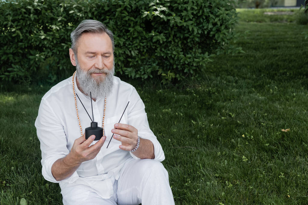 bearded healing guru holding diffuser and aroma sticks while sitting on lawn in garden - Photo, Image