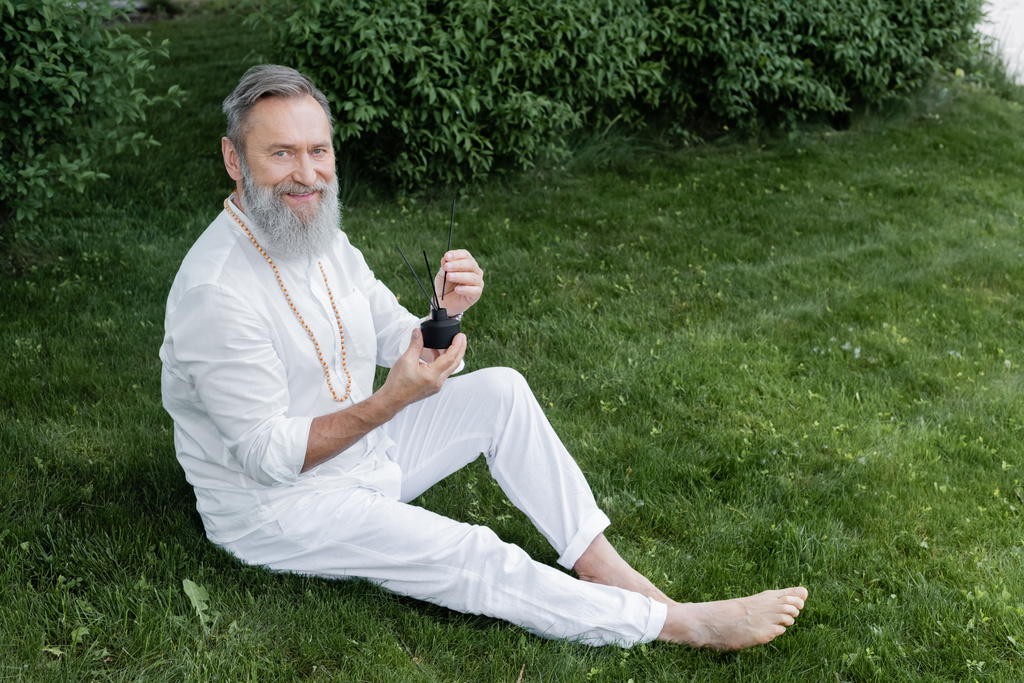 Glücklicher Senior-Yoga-Trainer sitzt mit Diffusor und Aromastäben auf grünem Rasen - Foto, Bild