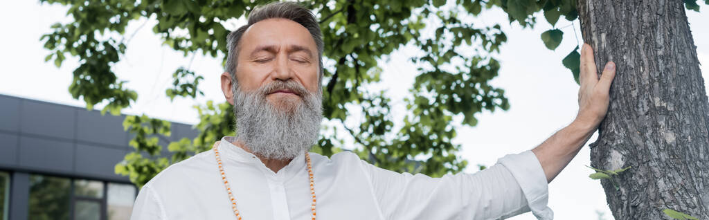 senior goeroe man met gesloten ogen mediteren in de buurt van boomstam, banner - Foto, afbeelding