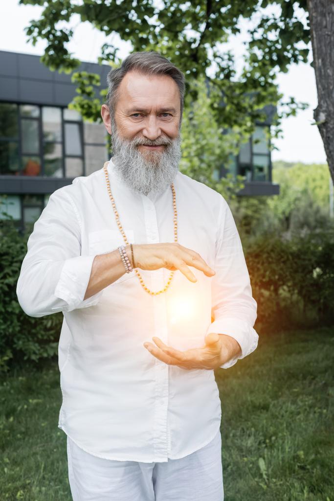 happy guru man looking at camera near shining aura between hands - Photo, Image