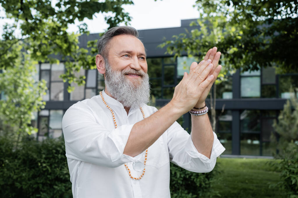 улыбающийся духовный наставник смотрит в сторону, медитируя с молитвенными руками в парке - Фото, изображение