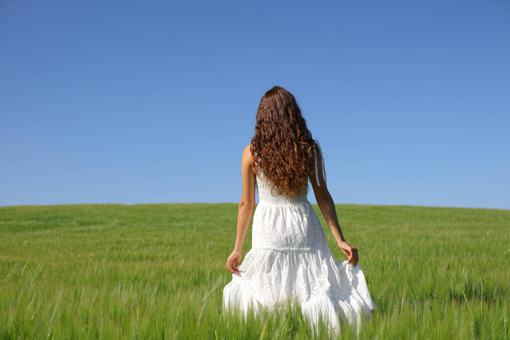Widok z tyłu kobiety w białej sukience spacerującej po zielonym polu pszenicy - Zdjęcie, obraz