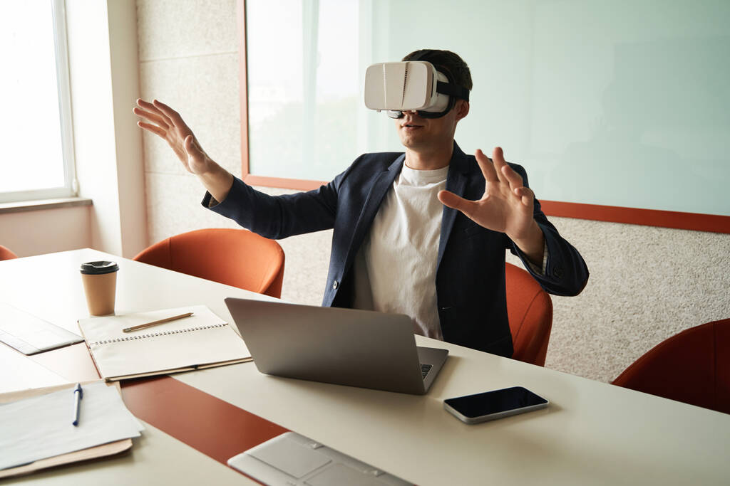 Travailleur de bureau au bureau devant un ordinateur portable assis au bureau et regardant à travers un casque de réalité augmentée - Photo, image