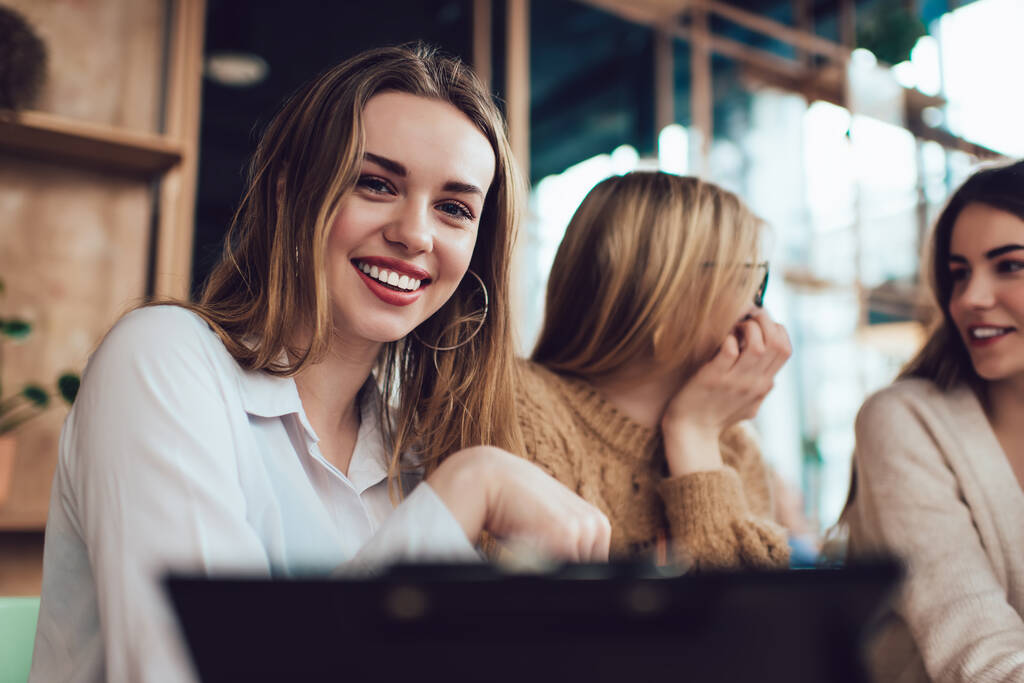 Mosolygós női szabadúszó alkalmi viselet írótáblával ül a modern kávézóban a barátaival, miközben együtt dolgoznak a távoli projekten - Fotó, kép