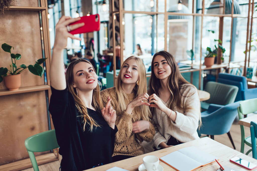 Çekici, gündelik giysiler içinde genç ve çekici kadınlar akıllı telefonlarıyla selfie çekerken aynı zamanda parlak zamanlı kafeden ödev yapıyorlar. - Fotoğraf, Görsel