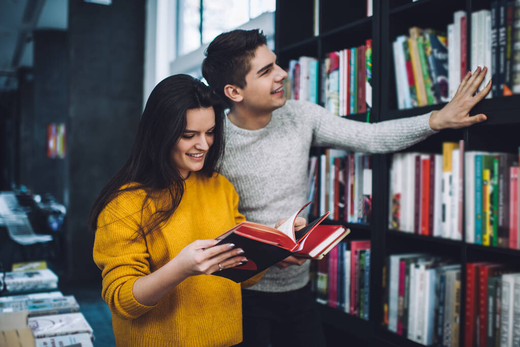 Homme optimiste regardant les noms des livres pendant le choix de la littérature à lire tout en souriant petite amie tourner des pages de livre examinant le contenu - Photo, image