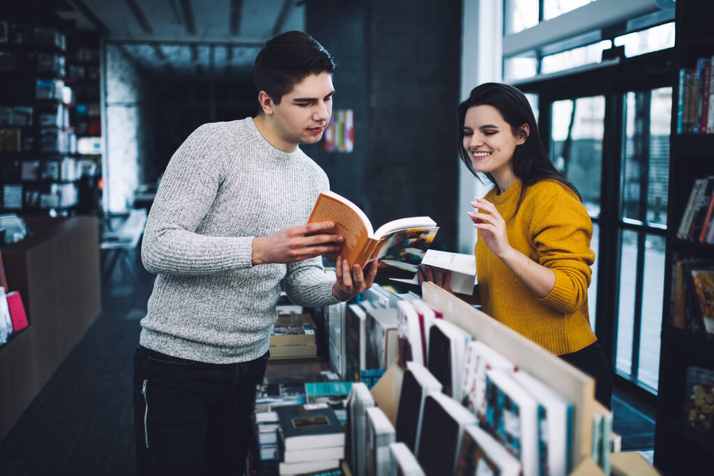 Konzentrierter Mann zeigt seiner lächelnden Freundin den Text eines offenen Buches, während er in der Nähe von Bücherregalen in einem geräumigen Geschäft steht und gemeinsam eine interessante Geschichte auswählt - Foto, Bild