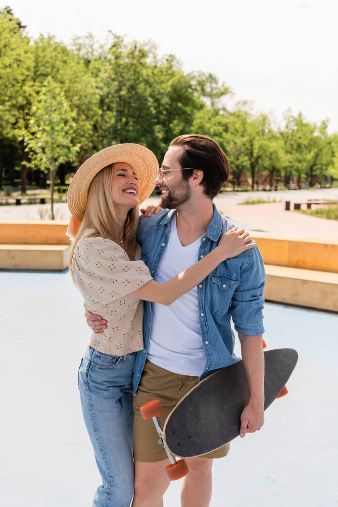 Woman in straw hat hugging boyfriend with longboard in skate park  - Photo, Image