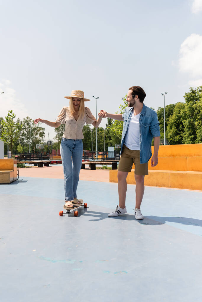Hombre sonriente en gafas de sol cogido de la mano de su novia montando longboard en skate park  - Foto, imagen